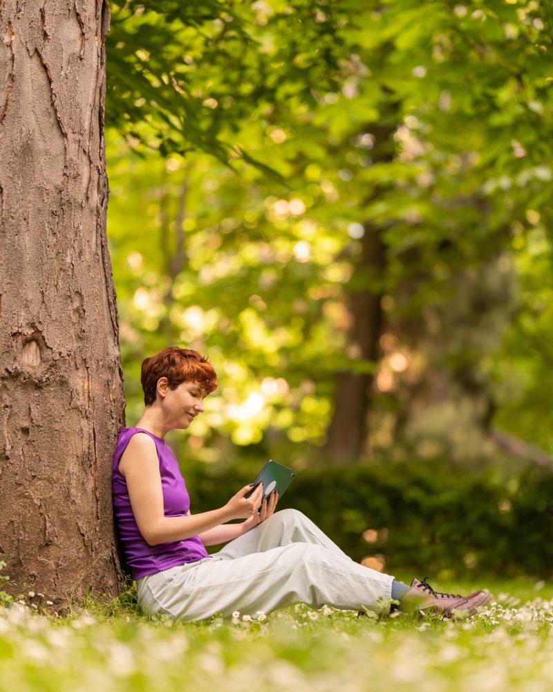 Side view of androgynous female in casual clothes sitting on lawn near tree and using tablet on sunny summer day in lush park