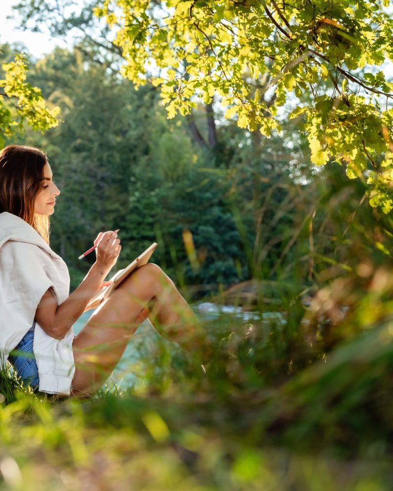 Young attractive girl writing and drawing in notebook sitting under the tree on nature in sunlight. Closeness to nature  inspires creativity. Urban escapism.